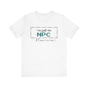 May include: A white short-sleeve t-shirt featuring a black speech bubble graphic. Inside the bubble, the text reads i'm just an NPC in a pixelated font, with the additional text [ press A to talk ] printed in a smaller font below the bubble.