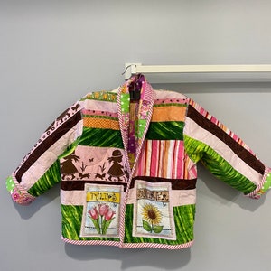 May include: A pink and green quilted jacket with a brown trim. The jacket has a patchwork design with different patterns and colors. The front of the jacket has two embroidered patches, one with the word "TULIPS" and a picture of tulips, and the other with the word "SUMMER" and a picture of a sunflower.
