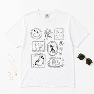 May include: A white short-sleeve t-shirt featuring a black line art graphic print of various dog breeds inside decorative frames, a bouquet of flowers, and small floral accents. The shirt has a crew neckline and a Comfort Colors brand label.