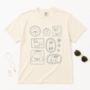 Könnte beinhalten: Ein cremefarbenes Kurzarm-T-Shirt mit einer schwarzen Strichgrafik verschiedener Hunderassen in dekorativen Rahmen, begleitet von Blumenillustrationen. Das T-Shirt hat einen Rundhalsausschnitt und ein Etikett am Kragen mit der Aufschrift Comfort Colors L-G-G.