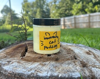 May include: A small glass jar with a black lid filled with a light yellow substance. The jar has a yellow label with black handwritten text that reads "banana cat pudding".