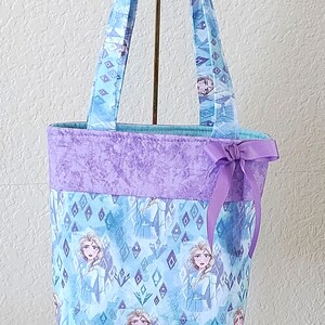 K&ouml;nnte beinhalten: Eine Tragetasche mit hellblauem Stoff, der mit einem Prinzessinnenmuster versehen ist. Der obere Teil ist einfarbig lavendelfarben, und eine lavendelfarbene Schleife ist angebracht. Die Tasche hat passende Griffe.