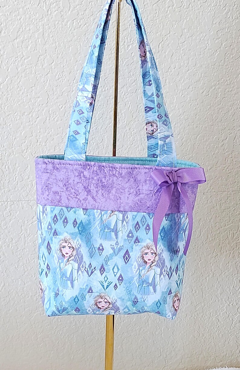 K&ouml;nnte beinhalten: Eine Tragetasche mit hellblauem Stoff, der mit einem Prinzessinnenmuster versehen ist. Der obere Teil ist einfarbig lavendelfarben, und eine lavendelfarbene Schleife ist angebracht. Die Tasche hat passende Griffe.