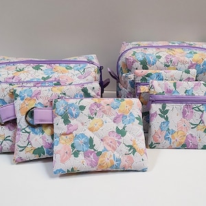 May include: A set of matching floral print bags in various sizes. The bags feature a white background with colorful flowers in shades of blue, yellow, and pink. Each bag has a purple zipper and trim. The set includes a large tote, a medium bag, and several smaller pouches.