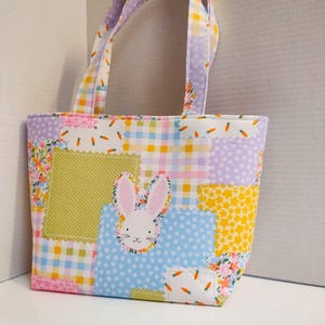 May include: A patchwork tote bag with a bunny applique. The bag features a variety of pastel fabrics, including gingham, floral, and polka dot patterns. The bunny has pink ears and a white face. The bag has two handles.