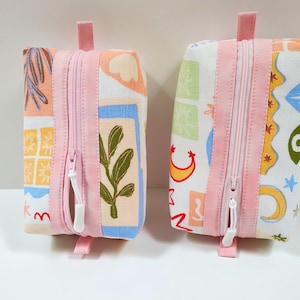May include: Two rectangular fabric pouches with pink zips and pull tabs. The pouches feature colourful patterns with botanical and abstract designs. The zips are centred, and the pull tabs are at the top. The pouches are on a white surface.
