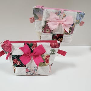 以下が含まれることがあります： 花柄パッチワーク模様のキルティングコスメティックバッグ2つ。各バッグには明るいピンクのリボンと、お揃いのピンクのジッパーが付いています。バッグの側面には小さなピンク色の布ループが付いています。バッグは白い表面にあります。