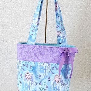 K&ouml;nnte beinhalten: Eine Tragetasche mit einem hellblauen und lila Design mit einer Figur. Die Tasche hat ein lila Band oben und eine lila Schleife an der Seite. Die Griffe sind hellblau.