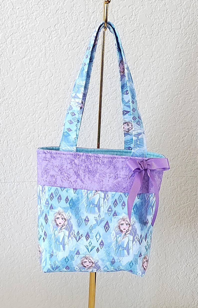 K&ouml;nnte beinhalten: Eine Tragetasche mit einem hellblauen und lila Design mit einer Figur. Die Tasche hat ein lila Band oben und eine lila Schleife an der Seite. Die Griffe sind hellblau.