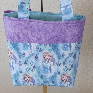 K&ouml;nnte beinhalten: Eine kleine Stoff-Tragetasche mit einem sich wiederholenden Muster einer Figur aus einem beliebten Animationsfilm. Die Tasche hat ein lila Oberband, einen blau-wei&szlig; gemusterten K&ouml;rper und passende Griffe. Der Innenraum ist aus hellblauem Stoff.