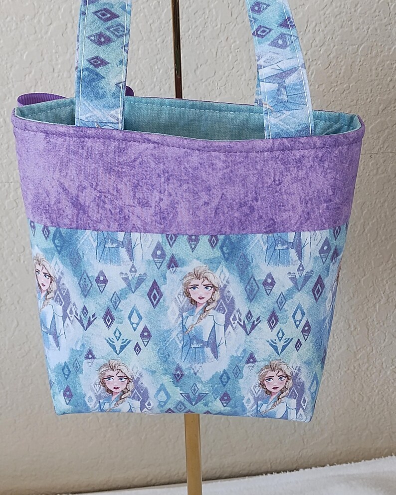 K&ouml;nnte beinhalten: Eine kleine Stoff-Tragetasche mit einem sich wiederholenden Muster einer Figur aus einem beliebten Animationsfilm. Die Tasche hat ein lila Oberband, einen blau-wei&szlig; gemusterten K&ouml;rper und passende Griffe. Der Innenraum ist aus hellblauem Stoff.