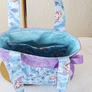 K&ouml;nnte beinhalten: Eine kleine Tragetasche aus hellblauem und lila Stoff mit einem sich wiederholenden Muster aus geometrischen Formen und einem Charaktergesicht. Die Tasche hat ein hellblaues Innenfutter und eine lila Schleife an der Seite.