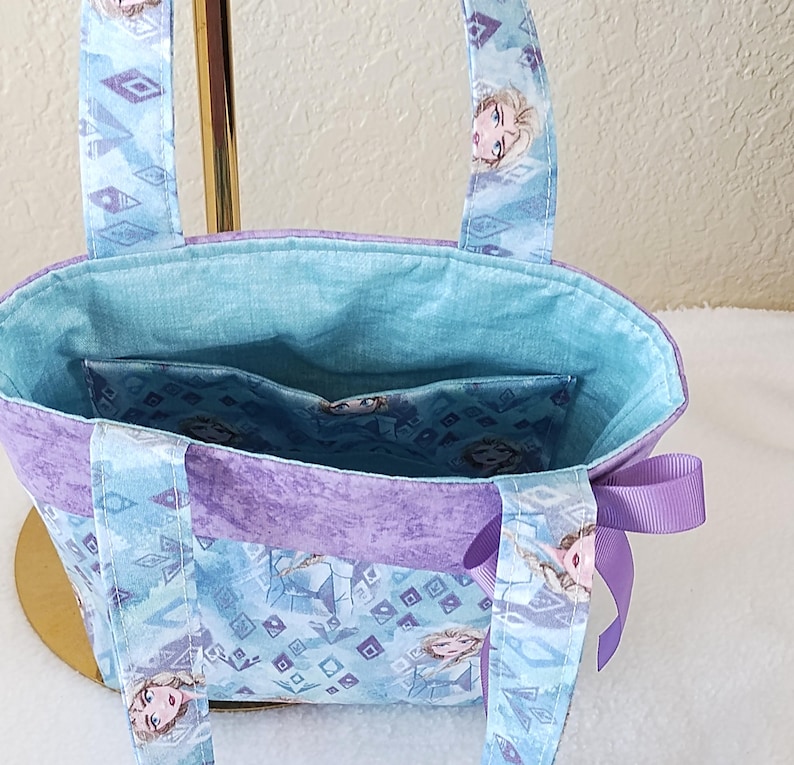 K&ouml;nnte beinhalten: Eine kleine Tragetasche aus hellblauem und lila Stoff mit einem sich wiederholenden Muster aus geometrischen Formen und einem Charaktergesicht. Die Tasche hat ein hellblaues Innenfutter und eine lila Schleife an der Seite.