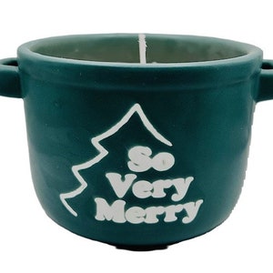 Könnte beinhalten: Ein tealgrüner Kerzenhalter aus Keramik in Form eines Topfes mit zwei Griffen. Der Kerzenhalter hat eine weiße Umrandung eines Weihnachtsbaums mit den Worten "So Very Merry" im Baum.