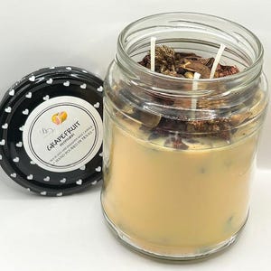 Könnte beinhalten: Eine Glas-Kerze in einem Einmachglas mit einem schwarzen und weißen Herzen-Muster auf dem Deckel. Die Kerze ist mit "Grapefruit" und "Putrify" beschriftet, sowie mit dem Text "28 oz Soy and Paraffin Wax Candle Hand Poured in Texas". Die Kerze hat ein helles orangefarbenes Wachs und getrocknete Blumen und Kräuter im Inneren.