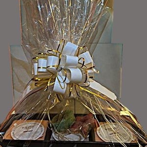 Könnte beinhalten: Ein Geschenkkorb, verpackt in durchsichtiger Cellophanfolie mit einer weißen Schleife und goldener Schleife. Der Korb enthält verschiedene Artikel, darunter ein Honigglas, eine Seife und eine Tüte Tee.