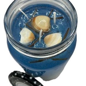 Könnte beinhalten: Eine blaue Glas-Kerze mit Muscheln und Dochten. Die Kerze befindet sich in einem durchsichtigen Glasgefäß mit einem schwarzen Deckel.