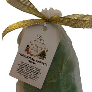 Könnte beinhalten: Ein klarer Cellophanbeutel, gebunden mit einem goldenen Band, enthält grüne, weihnachtsbaumförmige Seifen. Ein weißes Etikett mit dem Text "CHRISTMAS VARIETY SOAP" ist angebracht. Die Seife wiegt 79 Gramm.