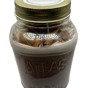 May include: A glass jar candle with a gold lid and a label that reads "Cinnamon Cinnamon Sticks Soy Wax Candle Hand Poured in Texas". The jar is labelled "Atlas Mason".
