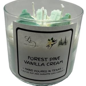 Könnte beinhalten: Eine klare Glaskerze mit einem Etikett mit der Aufschrift "Forest Pine Vanilla Cream". Die Kerze hat ein weißes und blaugrünes Farbschema mit dekorativen blaugrünen Akzenten oben. Die Kerze ist 629 Gramm.