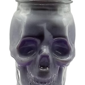Könnte beinhalten: Eine lila schädelformige Kerze in einem durchsichtigen Glas mit einem silbernen Metalldeckel. Die Kerze hat eine glatte, glänzende Oberfläche und ist so gestaltet, dass sie einem menschlichen Schädel ähnelt.