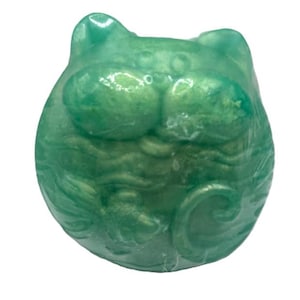 Könnte beinhalten: Eine grüne Keramik-Katzenfigur mit einem detaillierten Design. Die Katze hat einen runden Körper und ein breites, flaches Gesicht mit großen Augen.