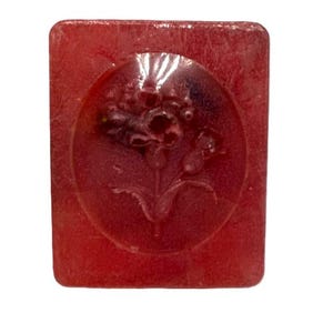 Könnte beinhalten: Eine rote, quadratische Seife mit einem geprägten Blumendesign in der Mitte.