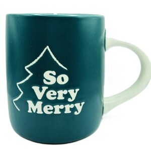 Könnte beinhalten: Eine tealblaue Keramiktasse mit einem weißen Henkel. Die Tasse hat einen weißen Umriss eines Weihnachtsbaums und den Text "So Very Merry" in Weiß.