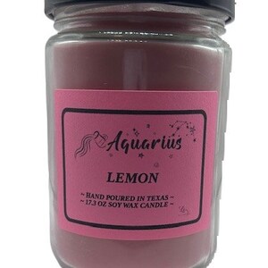 Könnte beinhalten: Eine rosa Glaskerze in einem Glas mit schwarzem Deckel. Das Etikett ist rosa mit schwarzem Text, der "Aquarius Lemon Hand Poured in Texas - 17.3 oz Soy Wax Candle - " lautet.