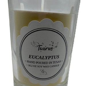 Könnte beinhalten: Eine durchsichtige Glaskerze in einem Glas mit einem weißen Etikett, auf dem "Taurus Eucalyptus - Hand Poured in Texas - 16.2 oz Soy Wax Candle" steht.