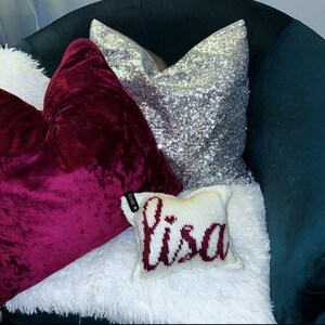 Puede incluir: Una colección de cojines decorativos en varias texturas y colores. Un cojín de terciopelo burdeos oscuro está junto a un cojín de lentejuelas plateadas. También hay una pequeña almohada de punto blanca con el nombre "Lisa" en escritura burdeos.