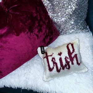 May include: A collection of decorative pillows in various textures and colors. A deep burgundy velvet pillow is next to a silver sequined pillow. A small, white knitted pillow with the name "Trish" in burgundy lettering is also present.