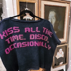 Puede incluir: Un suéter de punto azul marino oscuro con el texto "KISS ALL THE TIME. DISCO, OCCASIONALLY." bordado en hilo rosa brillante. La prenda tiene un cuello barco ancho y mangas cortas, exhibida en una percha negra sobre un fondo neutro.