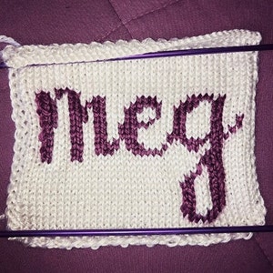 May include: A white knitted square with the name "Meg" in purple letters. The knitting needles are purple. The background is a solid purple color.