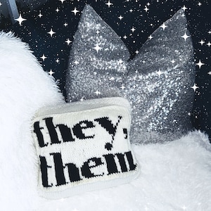 May include: A small, white knitted pillow with the words "they, them" in black lettering. The pillow is placed on a fluffy white surface, with a large, silver sequined pillow in the background. The background is a dark blue with white star accents.