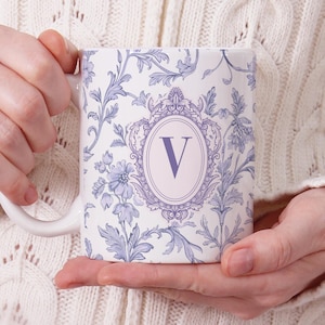 Peut inclure: Tasse en céramique blanche avec un motif floral dans les tons de bleu et de violet. La tasse porte la lettre "V" à l'intérieur d'un cadre ovale. La tasse est tenue par une personne portant un pull blanc en tricot torsadé.