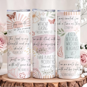 Puede incluir: Tres vasos de acero inoxidable blancos con citas inspiradoras y dise&ntilde;os florales y de mariposas. Los vasos presentan citas de los Salmos, Proverbios y otros vers&iacute;culos b&iacute;blicos. Los vasos est&aacute;n sobre una superficie de madera con flores en el fondo.