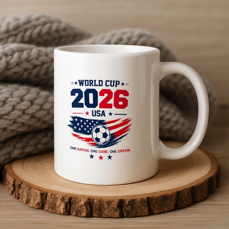 May include: A white ceramic mug featuring a graphic with the text WORLD CUP 2026 USA and ONE NATION. ONE GAME. ONE DREAM. The design includes a soccer ball centered over a stylized American flag motif in red, white, and blue with small stars.