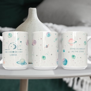 May include: Three white ceramic mugs with celestial designs. One mug features the Libra zodiac symbol and text. The other two mugs have a pattern of planets and stars. One mug has the text "Shine as bright as the stars."