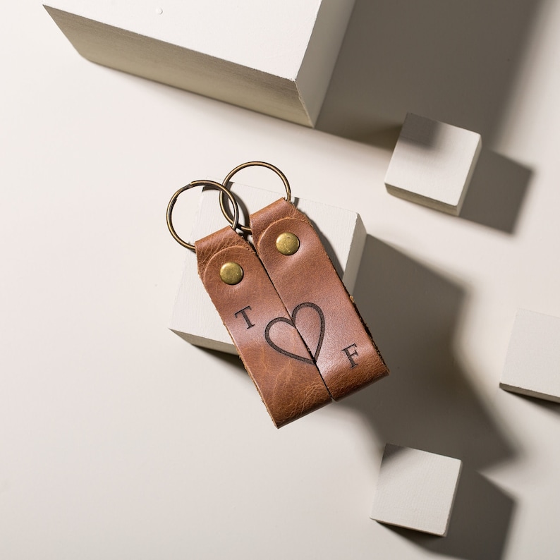 May include: Two brown leather keychains with gold rings. The keychains have the letters "T" and "F" engraved on them, with a heart shape between the letters.