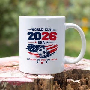May include: A white ceramic mug featuring a graphic with the text WORLD CUP 2026 USA and ONE NATION. ONE GAME. ONE DREAM. The design includes a soccer ball centered over a stylized American flag in red, white, and blue with small star accents.