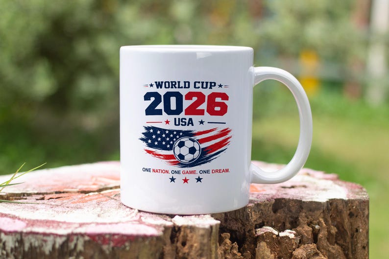 May include: A white ceramic mug featuring a graphic with the text WORLD CUP 2026 USA and ONE NATION. ONE GAME. ONE DREAM. The design includes a soccer ball centered over a stylized American flag in red, white, and blue with small star accents.