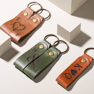 May include: Two sets of leather keychains with engraved initials and symbols. One set is brown with a heart and the initials "T & F". The other set is green with an ampersand and the initials "H & M". The third keychain is brown with a heart and the initials "K & O".
