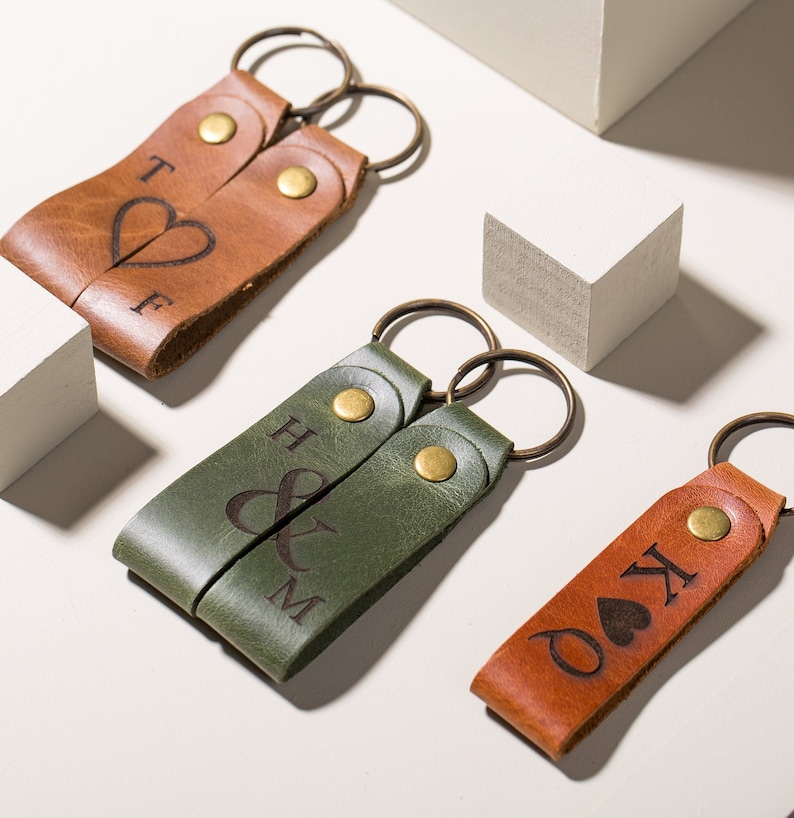 May include: Two sets of leather keychains with engraved initials and symbols. One set is brown with a heart and the initials "T & F". The other set is green with an ampersand and the initials "H & M". The third keychain is brown with a heart and the initials "K & O".