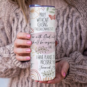 Puede incluir: Un vaso blanco y pastel con dise&ntilde;os florales y de mariposas, y texto inspirador. El texto incluye frases como "Orad sin cesar" y vers&iacute;culos b&iacute;blicos. El vaso es sostenido por una persona que lleva un su&eacute;ter beige.