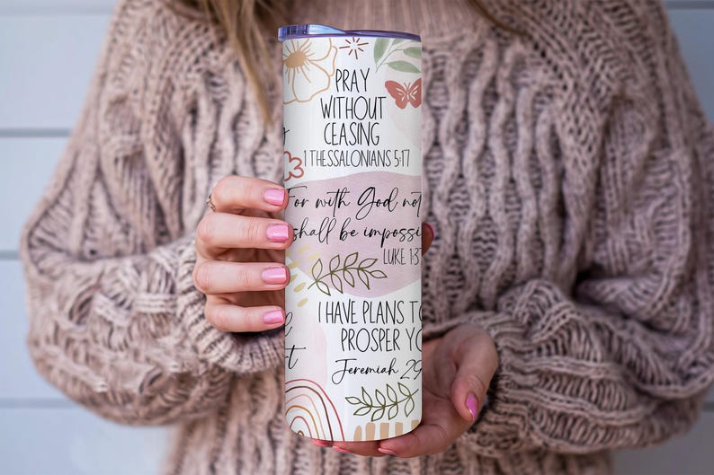Puede incluir: Un vaso blanco y pastel con dise&ntilde;os florales y de mariposas, y texto inspirador. El texto incluye frases como "Orad sin cesar" y vers&iacute;culos b&iacute;blicos. El vaso es sostenido por una persona que lleva un su&eacute;ter beige.