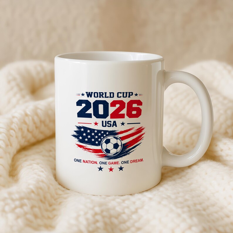 May include: A white ceramic mug featuring a graphic design for the 2026 World Cup in the USA. The design includes the text WORLD CUP 2026 USA, a soccer ball, a stylized American flag, and the text ONE NATION. ONE GAME. ONE DREAM. with stars.