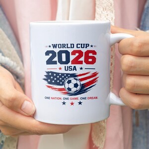 May include: A white ceramic mug featuring a World Cup 2026 USA graphic. The design includes the text 'WORLD CUP 2026 USA', a soccer ball over a stylized American flag, and the phrase 'ONE NATION. ONE GAME. ONE DREAM.' with red and blue stars.