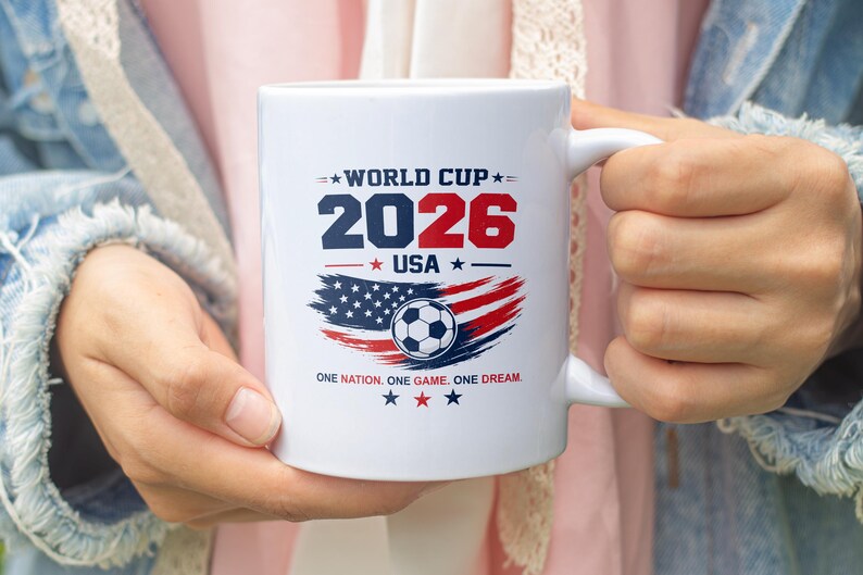 May include: A white ceramic mug featuring a World Cup 2026 USA graphic. The design includes the text 'WORLD CUP 2026 USA', a soccer ball over a stylized American flag, and the phrase 'ONE NATION. ONE GAME. ONE DREAM.' with red and blue stars.