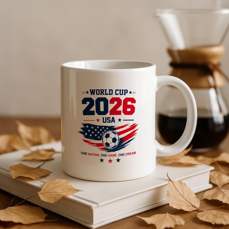 May include: A white ceramic mug featuring a graphic with the text WORLD CUP 2026 USA, a soccer ball, and a stylized American flag design. Below the graphic, the text reads ONE NATION. ONE GAME. ONE DREAM. The mug has a handle and a smooth finish.